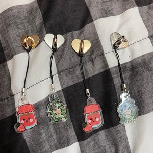 May include: Four phone charms with different designs. Each charm features a clear acrylic pendant with a cute illustration. The charms are attached to black cords with gold, silver, and rose gold heart-shaped hooks.
