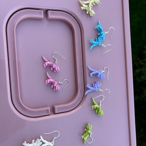 May include: A set of eight pairs of dinosaur earrings in various pastel colors, including pink, purple, blue, green, and yellow. The earrings are displayed on a pink surface.