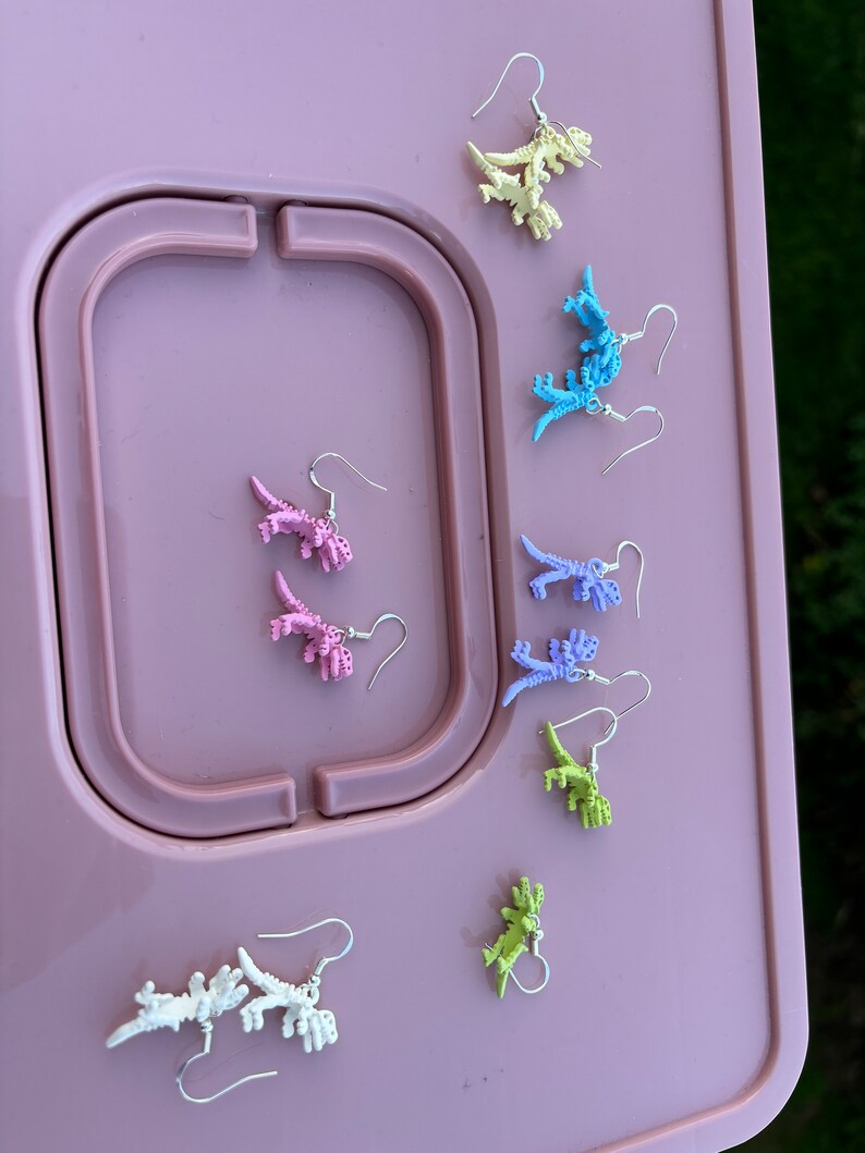 May include: A set of eight pairs of dinosaur earrings in various pastel colors, including pink, purple, blue, green, and yellow. The earrings are displayed on a pink surface.