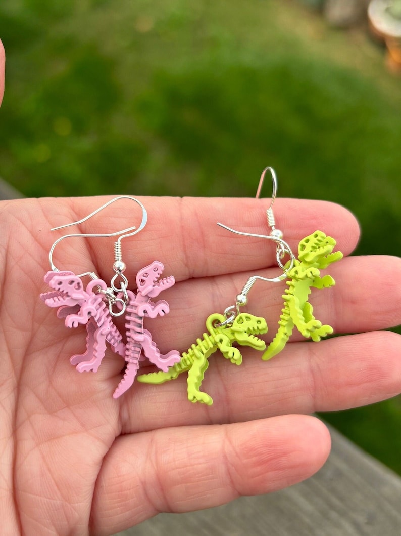 May include: A pair of earrings featuring dinosaur skeletons. One pair is pink and the other is green.
