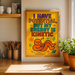 May include: A framed rectangular wall art print with a yellow background featuring the text I HAVE POTENTIAL, BUT MY ENERGY IS KINETIC in bold letters. Below the text is a cartoon illustration of an orange figure sleeping with zzz symbols.