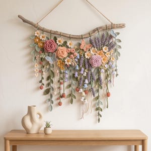 May include: A wall hanging featuring crocheted flowers, leaves, and butterflies in shades of pink, lavender, yellow, and green, suspended from a natural wooden branch with a string. The arrangement includes roses, daisies, and hanging vines.