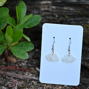 May include: A pair of white cloud-shaped earrings with silver hooks. The earrings are displayed on a white card.