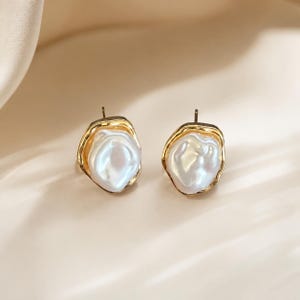 May include: A pair of stud earrings featuring irregular, lustrous white baroque pearls set within organic, textured gold-toned metal frames. The design highlights the natural, uneven shape of the pearls contrasted against the polished metallic edges.