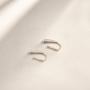 May include: A pair of silver-colored ear cuffs. Each cuff has a curved, elongated design with a rounded tip and a hook-like shape. The earrings are displayed on a light-colored surface.