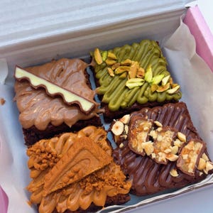 May include: A white cardboard box containing four square chocolate brownies with distinct toppings. One has light brown frosting and a chocolate piece, one has green pistachio cream and nuts, one has caramel spread and a biscuit, and one has chocolate drizzle and chopped nuts.