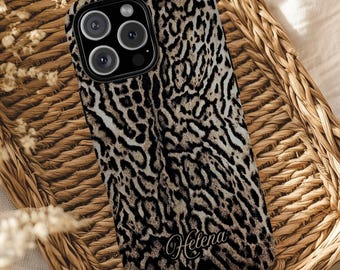 Può includere: Una custodia per telefono con un motivo leopardato nei toni del marrone, nero e crema. La custodia ha un ritaglio per la fotocamera e il nome "Helena" in un carattere corsivo. La custodia è appoggiata su un vassoio di vimini intrecciato.