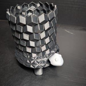 Pode incluir: Um vaso de plantas impresso em 3D em forma de tartaruga. O corpo tem um design geométrico cinza escuro, e a cabeça e as patas são cinza claro. O vaso tem um padrão hexagonal na parte superior.