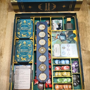 May include: An open board game box, titled "Wondrous Creatures", in teal and gold, filled with game components. The box contains cards, tokens, and game pieces in compartments. The game includes cards with the text "Players Turn" and "Tingent".