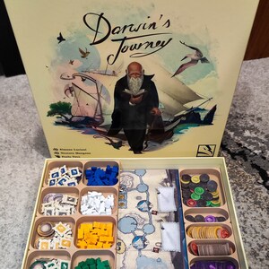 以下が含まれることがあります： 蓋が開いたベージュの箱で、中に「ダーウィンの旅」というボードゲームが入っています。ゲームボードと、さまざまなゲームピース、トークン、カードが見えます。箱の蓋には、本を持ったひげを生やした男性が船の上に立っているイラストが描かれています。