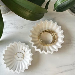 May include: Two white, fluted ceramic trinket dishes with gold jewellery. One dish holds a coiled gold bracelet, the other holds a pair of gold hoop earrings. The dishes are on a white marble surface, with green plant leaves in the background.