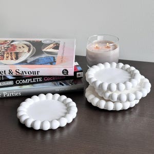 May include: Four white ceramic coasters with a raised, beaded edge. The coasters are stacked on top of each other.