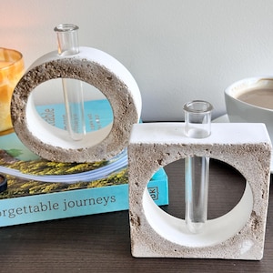 May include: Two white concrete bud vases, one round and one square, each with a clear glass tube for displaying flowers. The vases have a rough, textured exterior and a smooth interior. The round vase has a large circular opening.