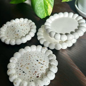 May include: Three white terrazzo coasters with a beaded edge. The coasters have a speckled pattern of brown, beige, and black. One coaster is stacked on top of two others. The coasters are on a dark wooden surface.