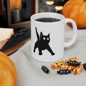 Może przedstawiać: Biały ceramiczny kubek z czarnym geometrycznym wzorem kota z świecącymi białymi oczami. Kubek zawiera ciemny płyn i jest otoczony dekoracjami na Halloween, w tym cukierkami i dyniami.