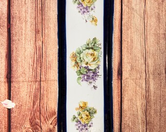 May include: A white ceramic serving dish with a blue border and floral design. The dish has two handles and is decorated with yellow roses and purple flowers.