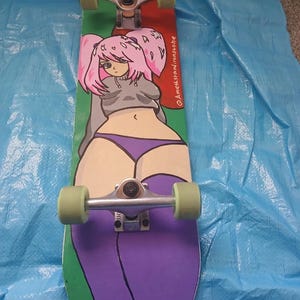 May include: A skateboard with a colorful design featuring a cartoon anime-style character with pink hair, a gray top, and purple shorts. The board has green and red accents, with green wheels and silver trucks. The text "@amethstardironsone" is visible.