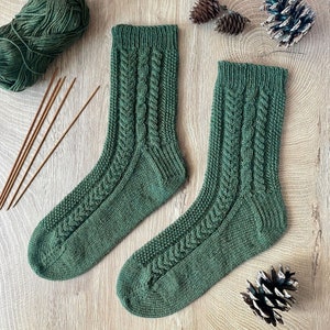 Pode incluir: Um par de meias verdes de malha com um design de cabo. As meias são exibidas em uma superfície de madeira com agulhas de tricô, fios e pinhas. As meias são de cor verde-média.