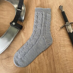 Può includere: Un calzino di lana grigio con un motivo a treccia. Il calzino è su una superficie di legno.