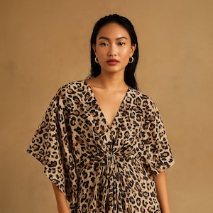 Op de afbeelding: Een lange, vloeiende jurk met een V-hals en korte mouwen. De jurk heeft een bruin en zwart luipaardprintpatroon. De stof is in de taille gerimpeld. Het model draagt gouden sieraden.