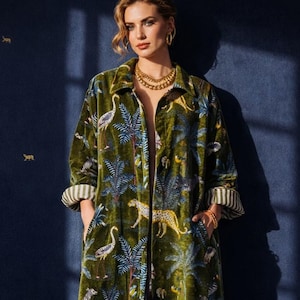 May include: An olive green velvet coat with a jungle pattern of palm trees, leopards, and birds. The coat has a relaxed fit, pockets, and striped cuffs. The model is wearing gold jewellery.
