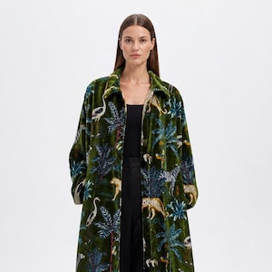 May include: A long, green velvet coat with a jungle pattern featuring leopards, zebras, and birds. The coat has a collared neckline and long sleeves. The model is wearing black trousers and a black top.