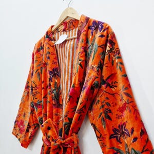 May include: An orange velvet robe with a floral pattern in shades of purple, green, and red. The robe has long sleeves and a matching tie belt. The interior lining features vertical orange and white stripes. The robe is hanging on a wooden hanger.