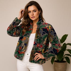 May include: A grey velvet jacket with a colourful floral design, including red, blue, and yellow flowers, and green leaves. The jacket has a high collar and is buttoned at the front. The model is wearing a white top and white trousers.