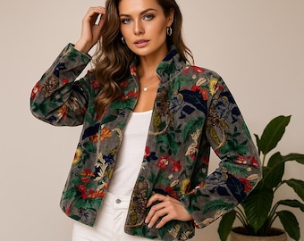 Op de afbeelding: Een grijze fluwelen jas met een levendig bloemenpatroon met rode, blauwe en gele bloemen en groene bladeren. De jas heeft een hoge kraag en is aan de voorkant dichtgeknoopt. Het model draagt een witte top en een witte broek.