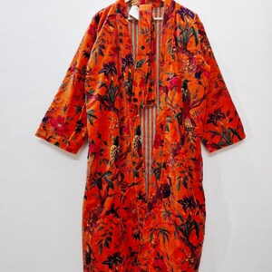 May include: An orange velvet robe with a floral and bird pattern. The robe is hanging on a wooden hanger. The pattern includes red, green, and purple accents. The robe has long sleeves and a tie closure.