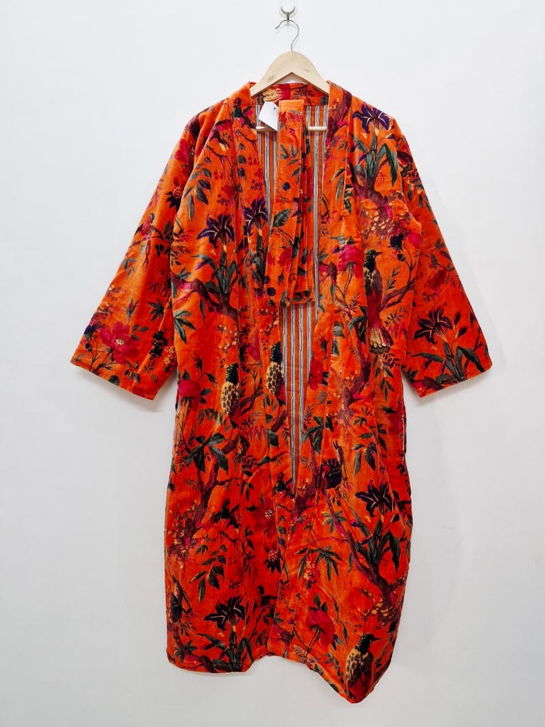 May include: An orange velvet robe with a floral and bird pattern. The robe is hanging on a wooden hanger. The pattern includes red, green, and purple accents. The robe has long sleeves and a tie closure.