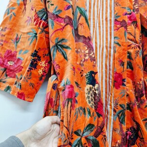 May include: An orange velvet kimono with a vibrant floral and bird print. The design features pink, red, and blue flowers, along with detailed illustrations of birds perched on branches. The interior has a vertical striped pattern.
