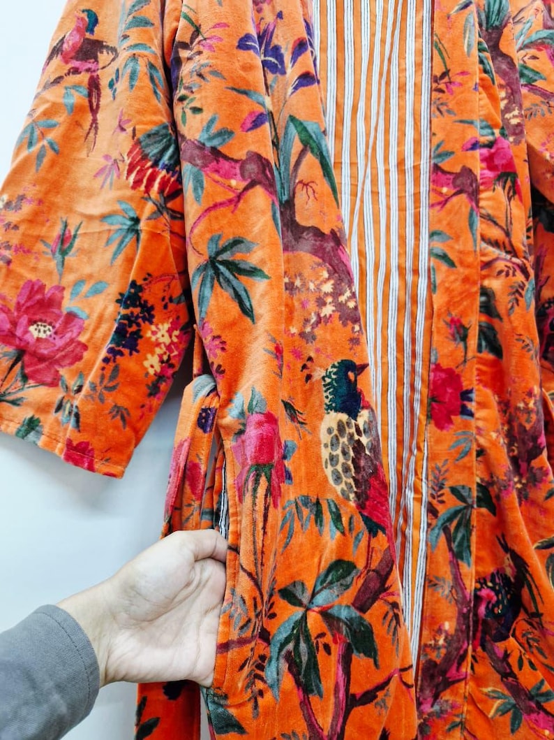 May include: An orange velvet kimono with a vibrant floral and bird print. The design features pink, red, and blue flowers, along with detailed illustrations of birds perched on branches. The interior has a vertical striped pattern.