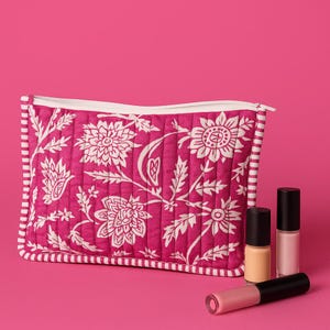 Puede incluir: Un neceser acolchado con estampado floral rosa con cremallera y ribete blancos. La bolsa está decorada con un estampado floral blanco. Tres frascos de esmalte de uñas y un tubo de brillo labial están junto a la bolsa sobre un fondo rosa.