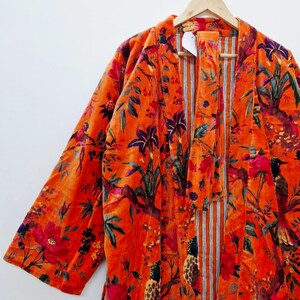 May include: A burnt orange velvet robe with a colourful floral and bird pattern. The robe has a striped inner lining and is suspended from a wooden hanger. The design incorporates red, purple, and green details.