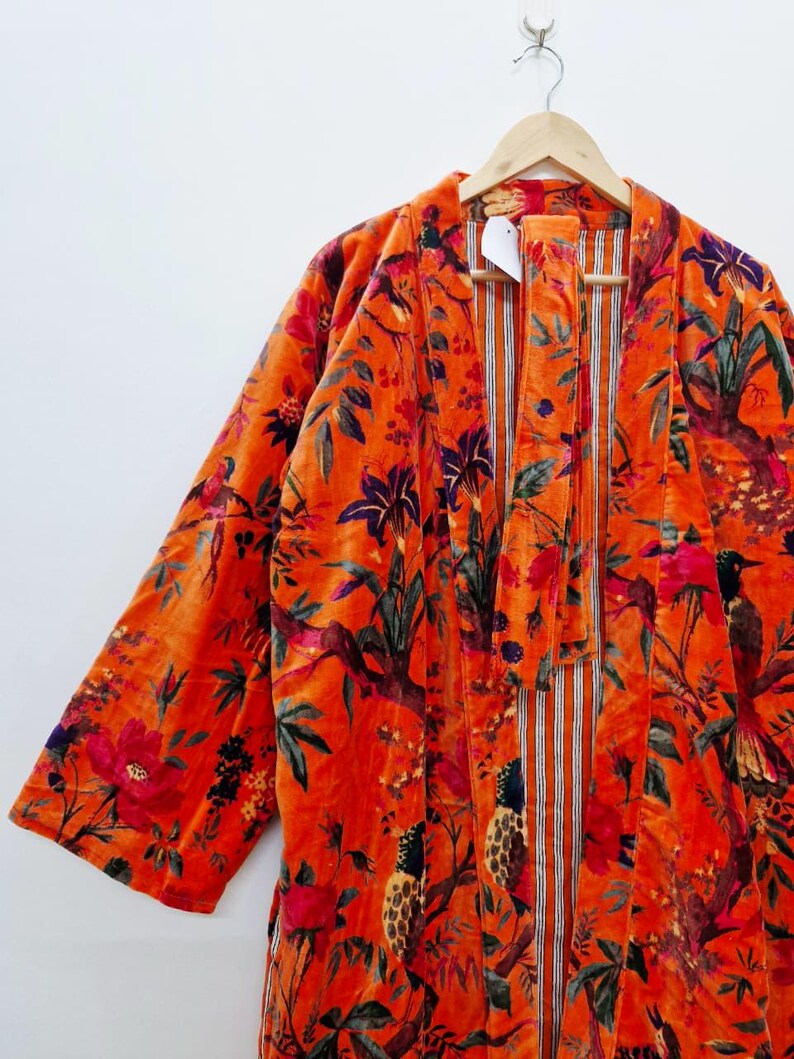 May include: A burnt orange velvet robe with a colourful floral and bird pattern. The robe has a striped inner lining and is suspended from a wooden hanger. The design incorporates red, purple, and green details.