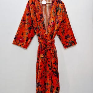 May include: An orange velvet robe with a floral design. The robe features long sleeves and a tie belt. The interior lining is striped. The robe is suspended from a wooden hanger.