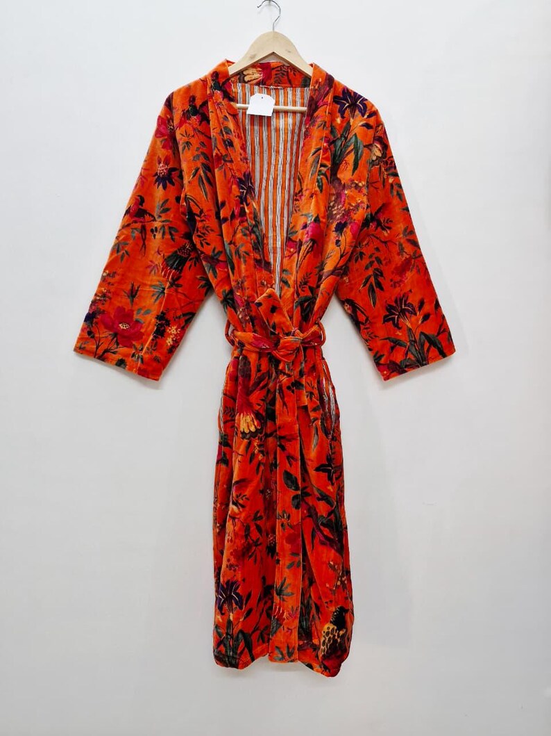 May include: An orange velvet robe with a floral design. The robe features long sleeves and a tie belt. The interior lining is striped. The robe is suspended from a wooden hanger.