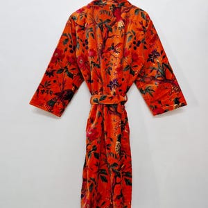May include: A vibrant orange velvet robe featuring a floral and bird design. The robe is suspended from a wooden hanger and includes a matching belt. The long-sleeved robe showcases a detailed botanical pattern in shades of red and green.