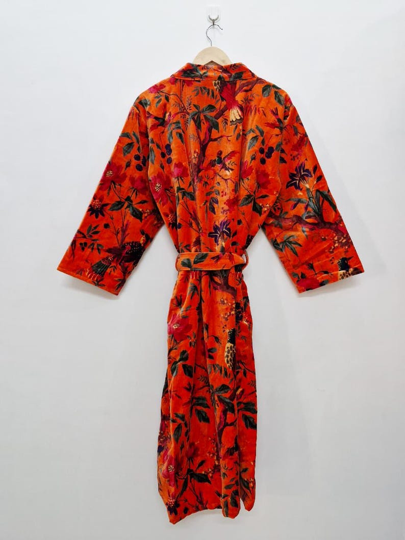 May include: A vibrant orange velvet robe featuring a floral and bird design. The robe is suspended from a wooden hanger and includes a matching belt. The long-sleeved robe showcases a detailed botanical pattern in shades of red and green.