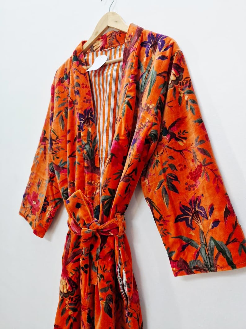 May include: An orange velvet robe with a floral pattern in shades of purple, green, and red. The robe has long sleeves and a matching tie belt. The interior lining features vertical orange and white stripes. The robe is hanging on a wooden hanger.
