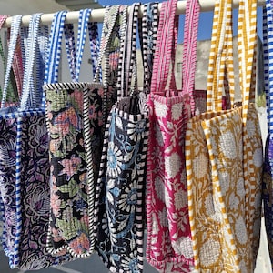 Könnte beinhalten: Eine Sammlung von gesteppten Tragetaschen, die in einer Reihe hängen, mit verschiedenen Blumenmustern in Schwarz, Pink und Gelb sowie kontrastierenden gestreiften Griffen und Besätzen in Blau, Weiß, Schwarz, Pink und Gelb mit detaillierten Nähten.
