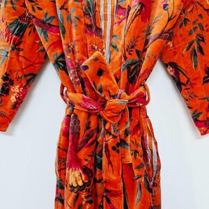 May include: An orange velvet robe with a floral design of birds and leaves. The robe has a tie belt and striped detailing on the inner collar and pockets. The sleeves are long, and the fabric looks soft and luxurious.