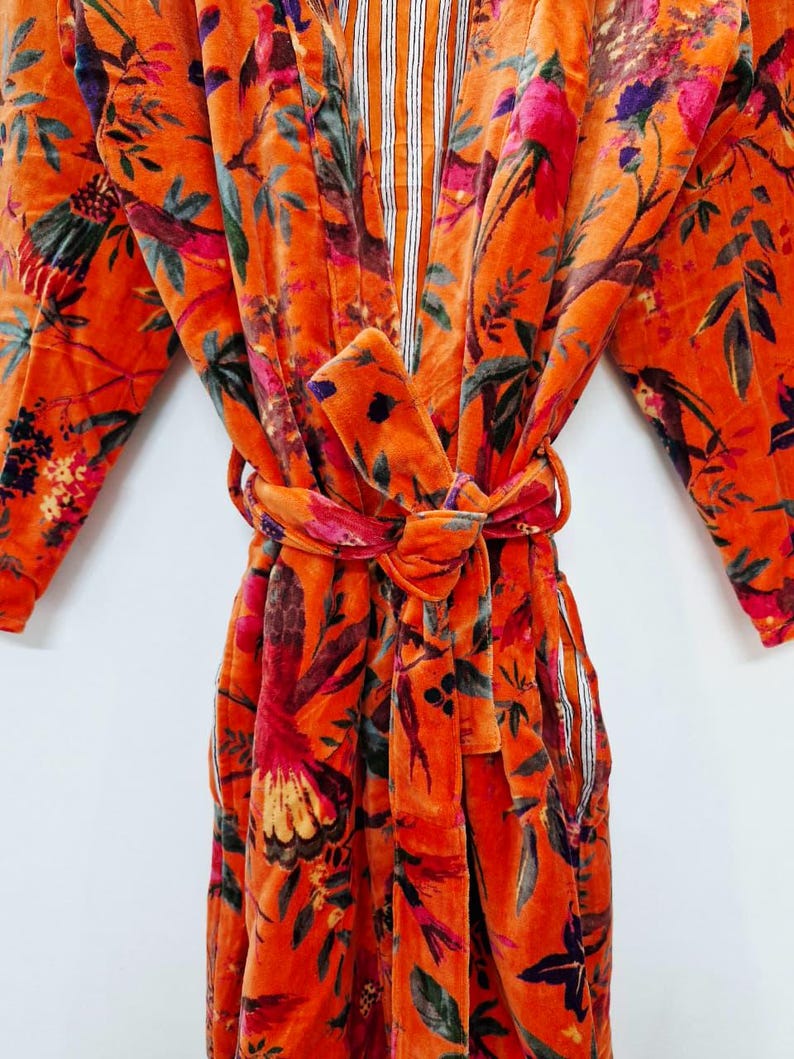 May include: An orange velvet robe with a floral design of birds and leaves. The robe has a tie belt and striped detailing on the inner collar and pockets. The sleeves are long, and the fabric looks soft and luxurious.