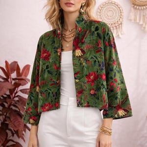 Könnte beinhalten: Grüne Samtjacke mit floralem Muster aus roten Blumen und Vögeln. Die Jacke hat einen hohen Kragen und Dreiviertelärmel. Das Model trägt die Jacke mit einem weißen Oberteil und einer weißen Hose.