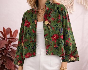 Op de afbeelding: Een groene fluwelen jas met een bloemenpatroon van rode bloemen en vogels. De jas heeft een hoge kraag en driekwart mouwen. Het model draagt de jas met een witte top en broek.