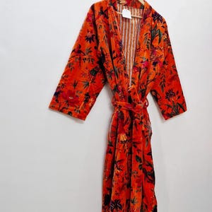 May include: A vibrant orange floral print robe with a matching belt, hanging on a wooden hanger. The robe features a pattern of red, green, and black flowers and foliage. The interior lining has vertical stripes.