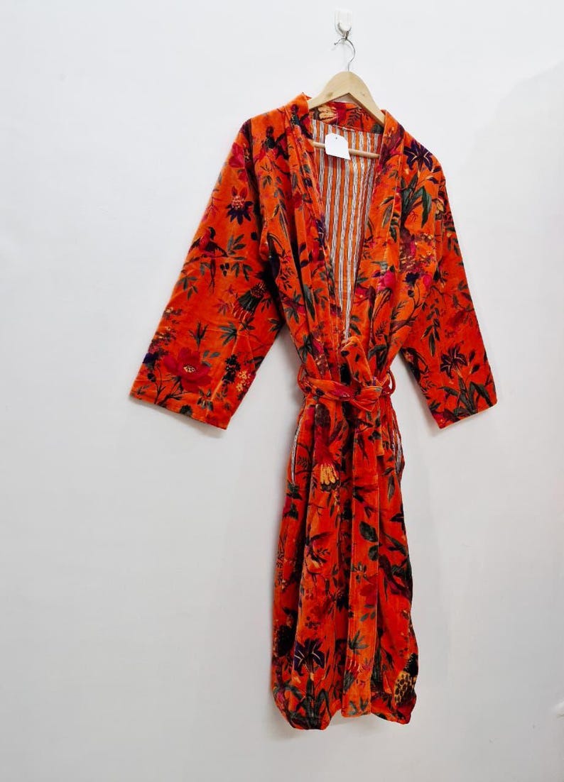 May include: A vibrant orange floral print robe with a matching belt, hanging on a wooden hanger. The robe features a pattern of red, green, and black flowers and foliage. The interior lining has vertical stripes.