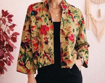 Op de afbeelding: Een jasje met bloemenprint met rode en groene accenten. De jas heeft een opstaande kraag en driekwartmouwen. Het model draagt een zwarte top en een zwarte broek. Gouden sieraden zijn zichtbaar.