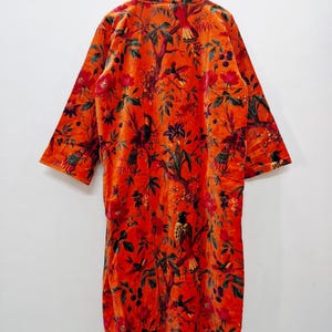 May include: An orange velvet robe with a vibrant floral and bird pattern. The long-sleeved robe features a detailed design of flowers, leaves, and birds in shades of red, green, and black. The robe is hanging on a wooden hanger.